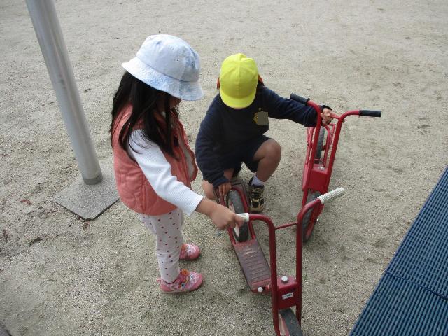 幼稚園の友達が保育所の友達にスクーターの使い方をやさしく教えてあげています。