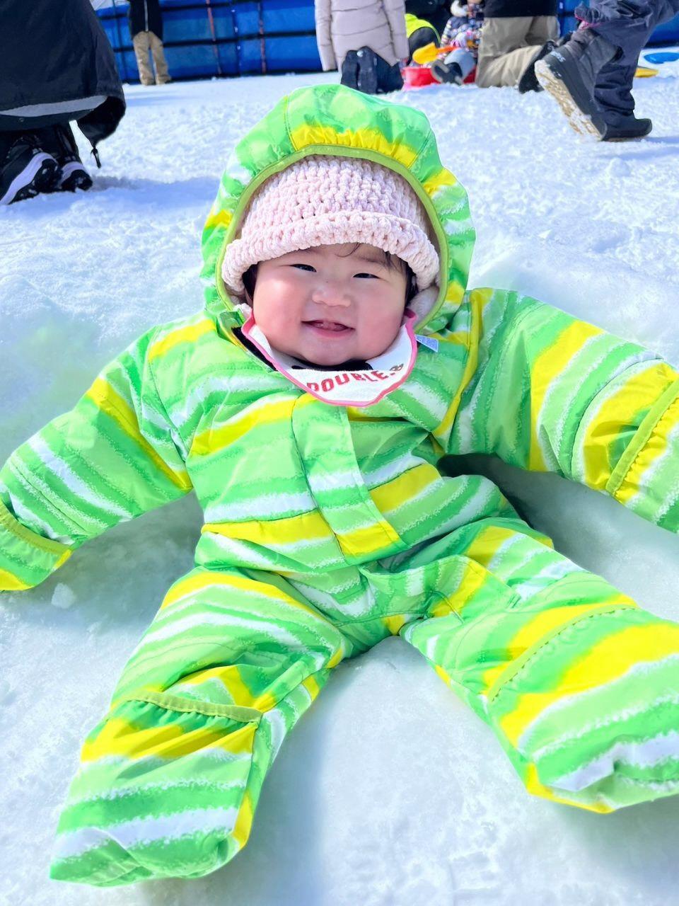 雪の上で大の字で座っている蛍光緑の服を着た赤ちゃんの写真
