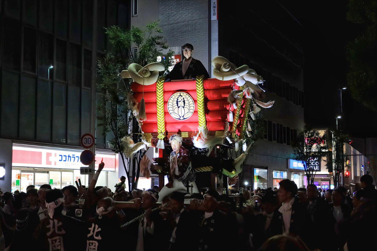 人が上に座っている布団太鼓を多くの人が囲んでいる写真